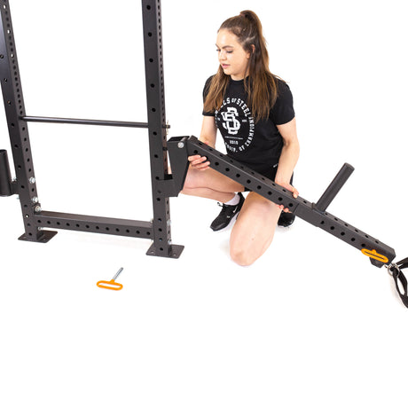 A woman in a black T-shirt and shorts kneels on the white floor, adjusting the Bells of Steel Belt Squat / Lever Arms Rack Attachment with focus, a small orange handle nearby.