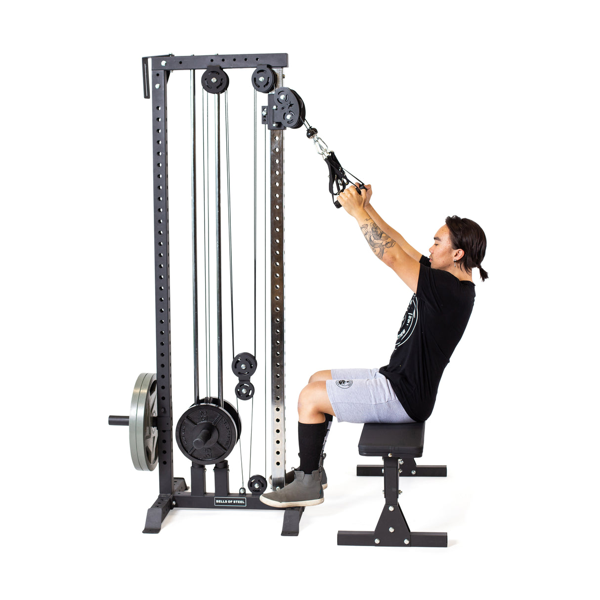 Using the Bells of Steel Cable Tower in their home gym, a person exercises by pulling down the handles for an effective strength workout. They are wearing a black shirt, gray shorts, and black shoes while using the multi-pulley system with a weighted stack.