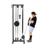 A person in a black t-shirt and gray shorts works out at home with the Cable Tower from Bells of Steel, skillfully pulling a handle on this advanced machine with multiple pulleys and weight plates.