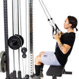 A person with long hair sits on a workout bench in their home gym using the Bells of Steel Cable Tower. Dressed in a black shirt and gray shorts, they pull the handles attached to the cables for exercises.