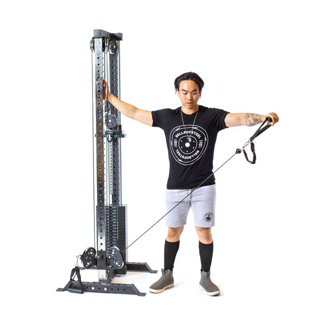 A person exercises on the Bells of Steel Cable Tower, pulling a handle with one arm extended to the side. They wear a black T-shirt, gray shorts, and knee-high black socks in their home gym. The background is white, and weights are loaded on the machine.