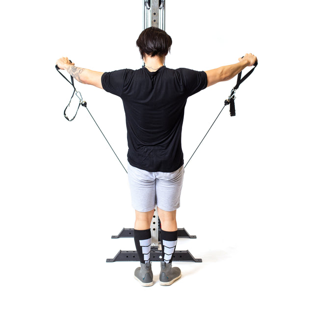 Using the Bells of Steel Cable Tower, a person enhances their home gym workout by pulling two handles outward. They're in a black shirt, gray shorts, and striped socks against a white background.