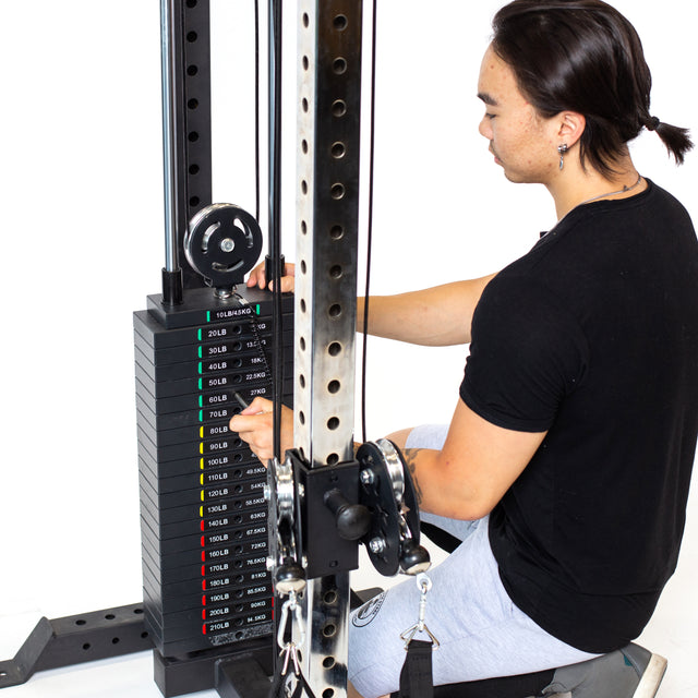 A person in a black shirt and gray pants adjusts the weight on the Bells of Steel Cable Tower. The stack shows weights from 20 lbs to 200 lbs. With their back to the camera, the individual uses this machine, ideal for home gyms, set against a white background.