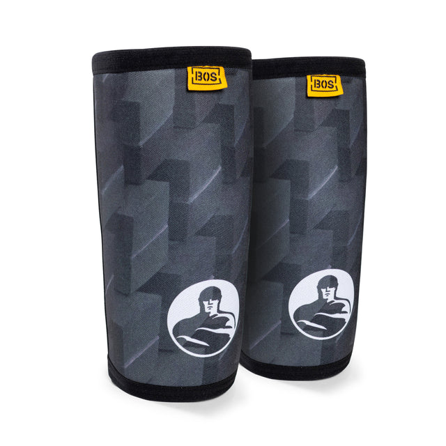 A pair of black and gray elbow sleeves showcasing a geometric design and a logo with a stylized figure. Engineered for compression and support, the top edge includes a small yellow tag displaying the brand name, Bells of Steel. They are positioned against a plain white background.