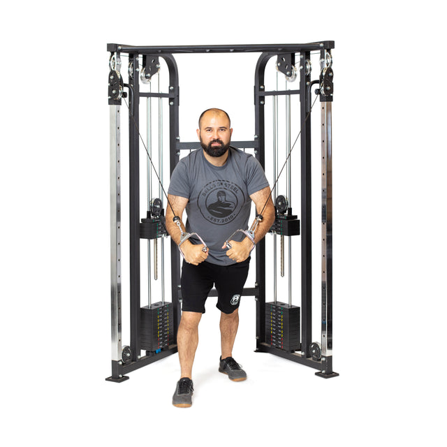 A bearded man exercises with focus on a Bells of Steel Functional Trainer. In a gray T-shirt and black shorts, he skillfully pulls the cables to his chest against a plain white background, highlighting the sleek design of the weight stacks and equipment.