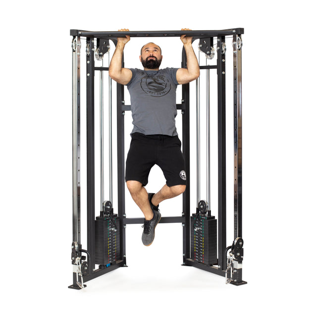 A bearded man skillfully performs a pull-up on the Bells of Steel Functional Trainer. Wearing a gray t-shirt, black shorts, and black shoes, he confidently maneuvers among the weight stacks and pulleys of the cable workout machine.