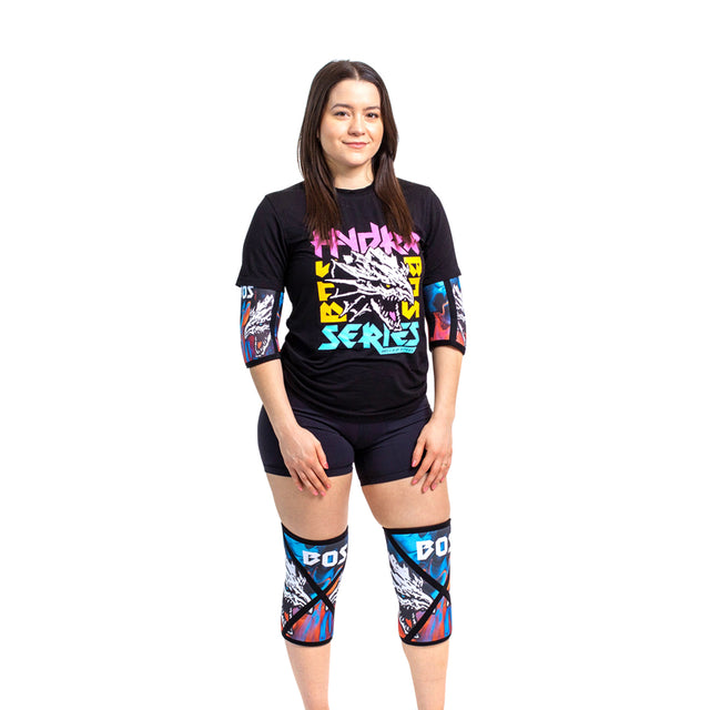 A person with long hair stands confidently in a black T-shirt featuring colorful graphics, paired with black shorts. They wear vibrant Bells of Steel Knee Sleeves, which provide compression and support, displaying a bold design against the plain white background.