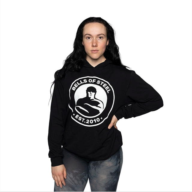 A person with long black hair stands against a white background, wearing the buttery soft Bells of Steel Bamboo Hoodie, featuring an emblem of a weightlifter and "Est. 2010" text, paired with gray camouflage leggings. This sustainable choice from Bells of Steel is both stylish and eco-friendly.