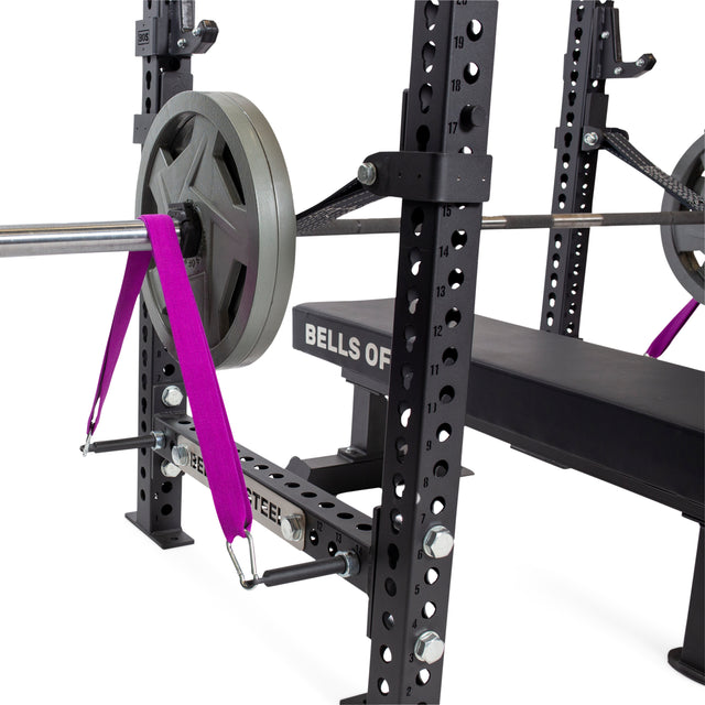 A gym bench press setup showcasing Bells of Steel's Band Pegs with Carabiners, featuring a barbell loaded with weight plates, secured by distinctive purple resistance bands and complemented by black equipment with numbered holes and a padded bench underneath to ensure stability and versatility for your workouts.