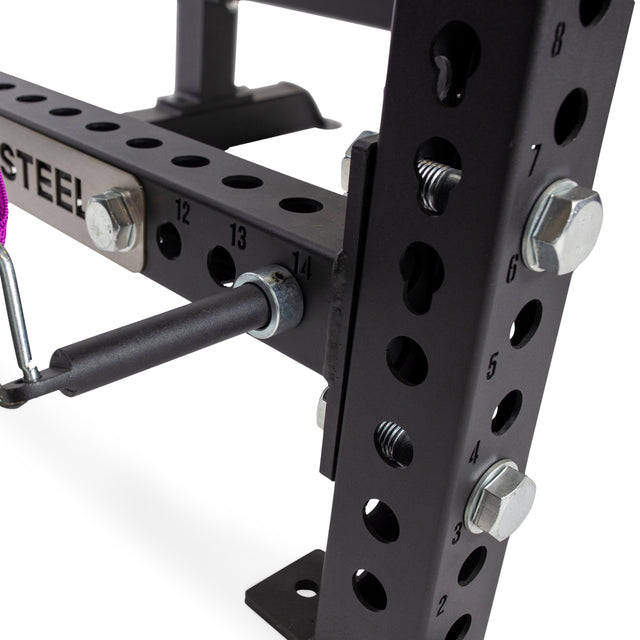 Close-up of a black metal exercise rack featuring numbered holes for adjustable settings, with a pin inserted into hole 14. The frame is secured with large bolts, and Band Pegs with Carabiners from Bells of Steel are attached. A partial logo displaying the word "STEEL" can be seen.