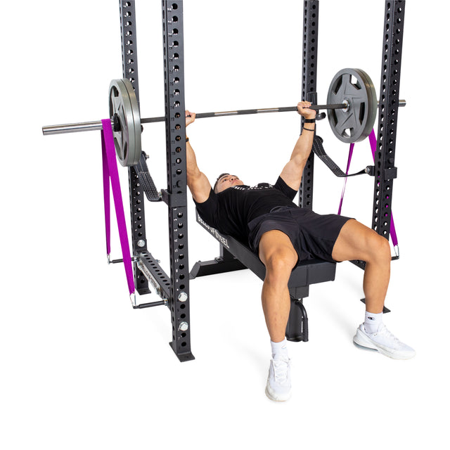 A person is executing a bench press with a barbell, incorporating resistance bands for extra tension. They are positioned on a weight bench situated inside a power rack and are using the Bells of Steel Band Pegs with Carabiners to secure the bands. The individual is dressed in a black shirt, black shorts, and white shoes.