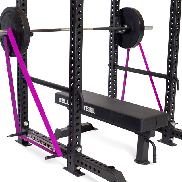 A Bells of Steel weightlifting bench includes a barbell set on a rack with weight plates on each side. For added support or resistance, it features purple Standard Band Pegs attached to the power rack.