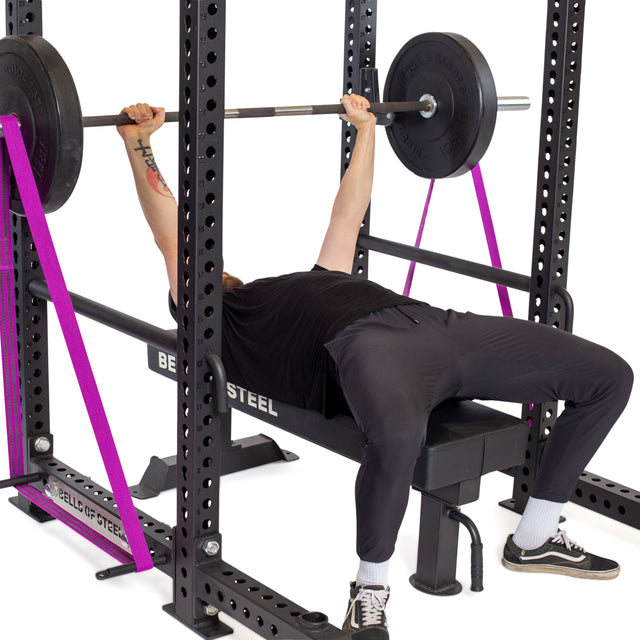 A person is executing a bench press on a power rack, utilizing the Bells of Steel Standard Band Pegs with pink resistance bands for added intensity. Dressed in a black shirt, dark pants, and sneakers, they skillfully handle the barbell and weight plates.