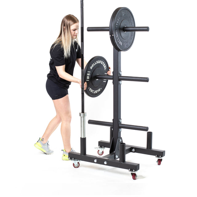 Dressed in black with yellow sneakers, a person adjusts a barbell on the Bells of Steel Bumper Plate Weight Tree and Bar Holder. The equipment has lockable wheels for easy mobility and stands against a plain white background.