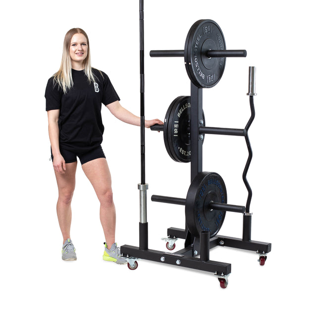 A person in athletic wear smiles beside a Bumper Plate Weight Tree And Bar Holder from Bells of Steel, with assorted barbells and kettlebells arranged neatly on the lockable wheel storage system against a plain white background.