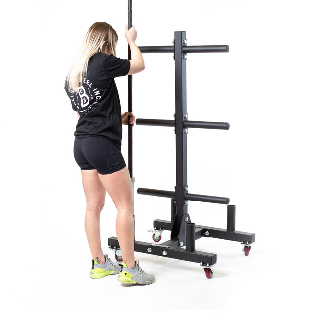 A person with long hair adjusts the Bells of Steel Bumper Plate Weight Tree and Bar Holder on lockable wheels, wearing a black shirt, shorts, and grey shoes with yellow accents against a plain white background.