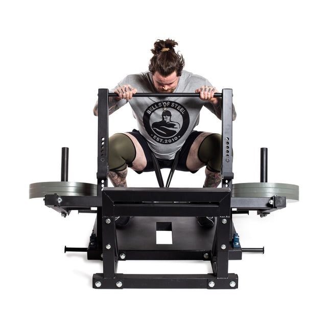 A person with a top knot uses the Bells of Steel Belt Squat Machine, grasping it tightly. They wear a gray t-shirt with a circular logo, black shorts, and knee pads. This setup is ideal for building lower body strength in their home gym, featuring weights on both sides and a sturdy frame.