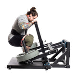 A tattooed man wearing a knee sleeve performs squats on the Bells of Steel Belt Squat Machine, loaded with weight plates, showcasing lower body strength training in a home gym setup against a plain white background.