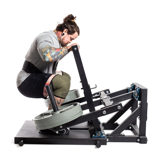 A tattooed man wearing a knee sleeve performs squats on the Bells of Steel Belt Squat Machine, loaded with weight plates, showcasing lower body strength training in a home gym setup against a plain white background.
