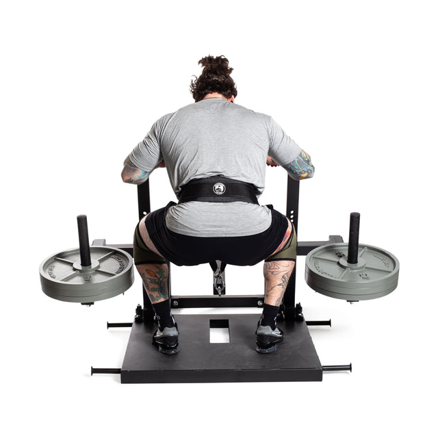 A person showcases impressive lower body strength on a Belt Squat Machine by Bells of Steel. Sporting tattoos and dressed in a gray shirt, black shorts, and a weightlifting belt, they power through weighted plate resistance in their home gym.