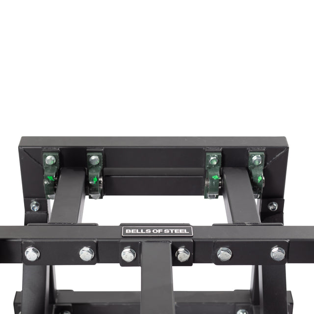 A close-up of the Belt Squat Machine's sturdy black metal frame by Bells of Steel shows green rollers and bolts. Ideal for boosting lower body strength at home, the industrial design prominently displays the brand label in the center.