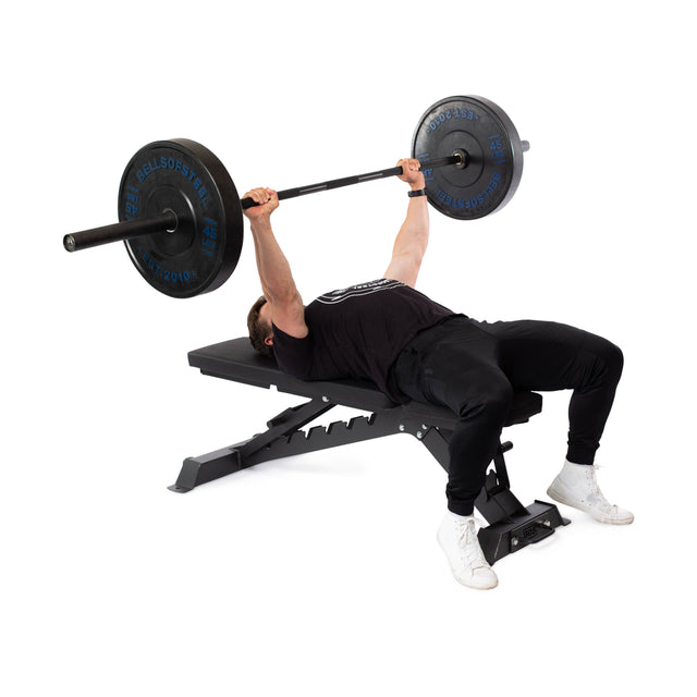 A person on the Bells of Steel Buzz-Saw Heavy-Duty Adjustable Bench lifts a barbell with weights above their chest, dressed in a black shirt, black pants, and white shoes. The setup ensures they can easily handle up to the max capacity effortlessly.