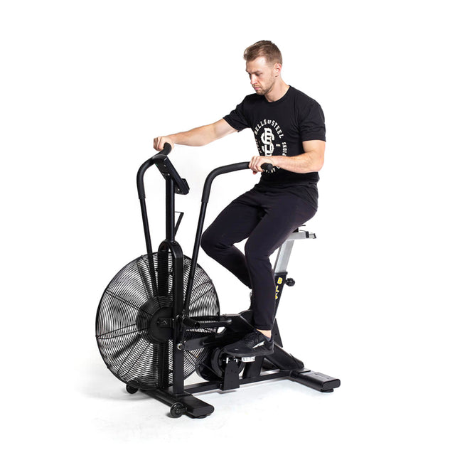 A man in black workout clothes vigorously exercises on a Bells of Steel Blitz Air Bike, an ideal home gym addition, as the large fan wheel hums against the plain white background, delivering an effective full-body workout.