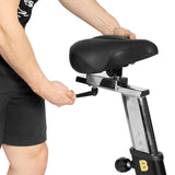 A user adjusts the seat height of the Bells of Steel Blitz Air Bike with a side knob. The bike seat and part of their arm and shorts are visible—ideal for customizing full-body home gym workouts.