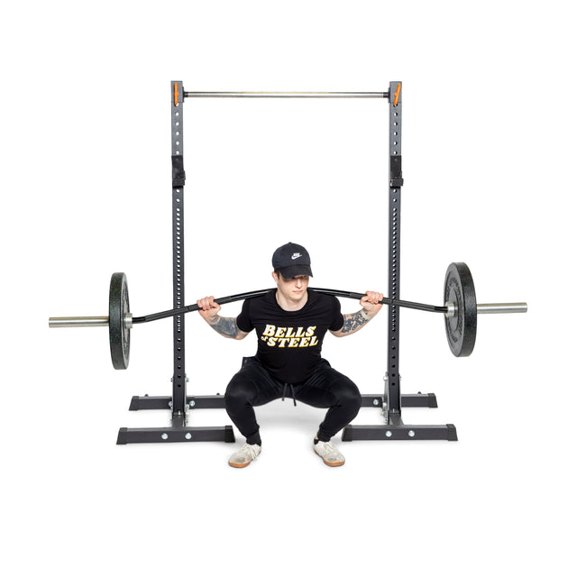 Dressed in all black and a "Bells of Steel" T-shirt, the person performs a squat with a Curved Squat Bar on their shoulders. Standing in front of a squat rack against a white backdrop, their gray shoes provide stability as they focus on relieving stress and reducing joint strain.