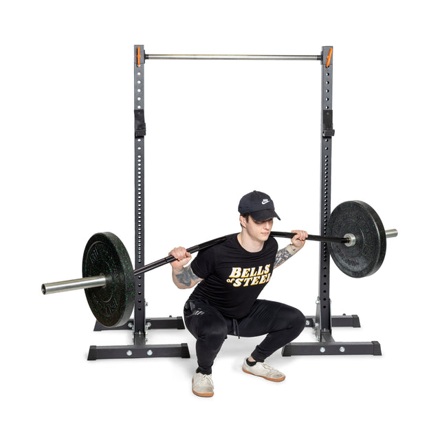 A person in a cap, black shirt, and athletic pants performs a barbell squat in a squat rack. The shirt says "BELLS OF STEEL." Using the Curved Squat Bar by Bells of Steel, they are in a low squat position on a white background to alleviate stress and reduce joint strain.