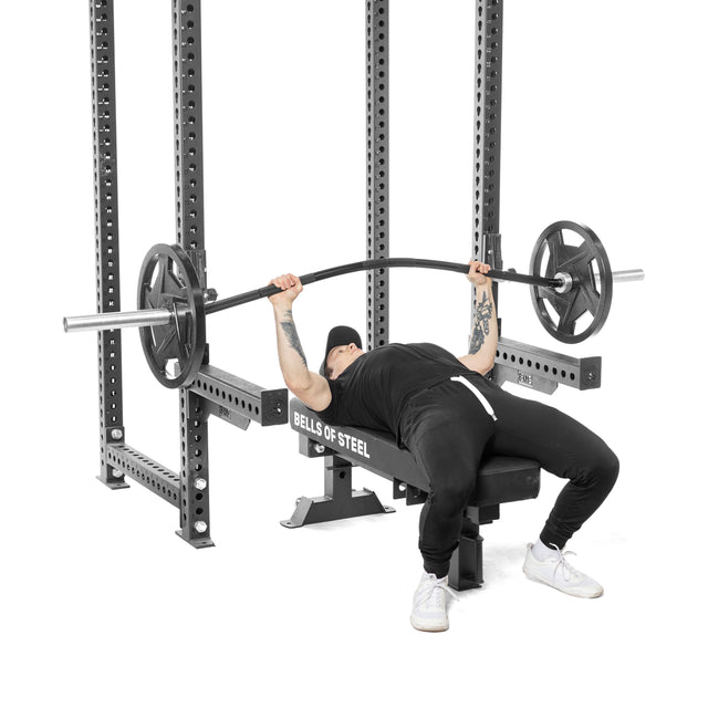 A person in black athletic wear and a cap bench presses with a Bells of Steel Curved Squat Bar, designed to reduce joint stress. They lie on a Bells of Steel bench, gripping the bar loaded with weights within a squat rack.