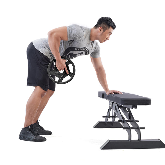 A man performs a bent-over row using a Black Mighty Grip Olympic Weight Plate from Bells of Steel in one hand, supporting himself with the other on a workout bench. Dressed in a gray t-shirt, black shorts, and black sneakers, he stands out against the plain white background.