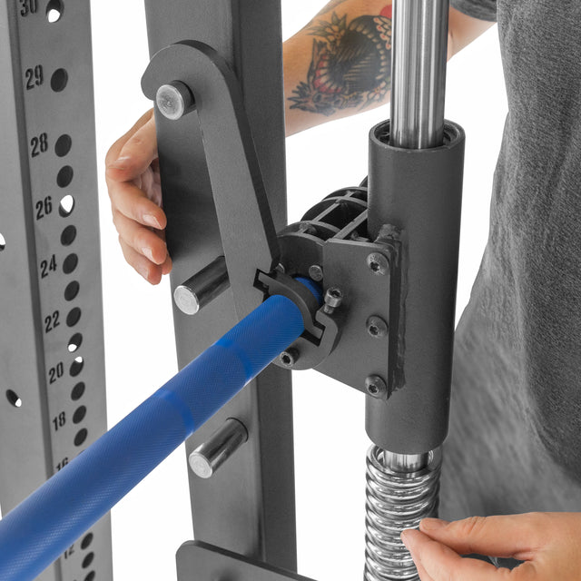 A person adjusts the blue lever on a Bells of Steel Smith Machine Rack Attachment, securing the mechanism and safety lock while a colorful tattoo is revealed on their arm. Nearby, metal rods and numbered holes highlight the equipment's versatility.