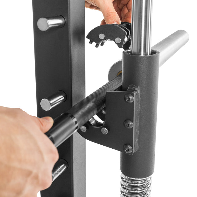 Close-up of hands securing a black barbell with a safety latch on the Bells of Steel Smith Machine Rack Attachment, featuring metal rods and springs in a gym setting.