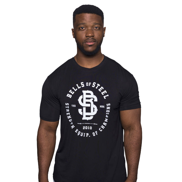 A man stands against a plain white background, showcasing the comfort and style of the Bamboo T-Shirt by Bells of Steel. The black workout shirt features "Bells of Steel Strength Equip. of Champions" and a central white logo, highlighting its sustainable design.