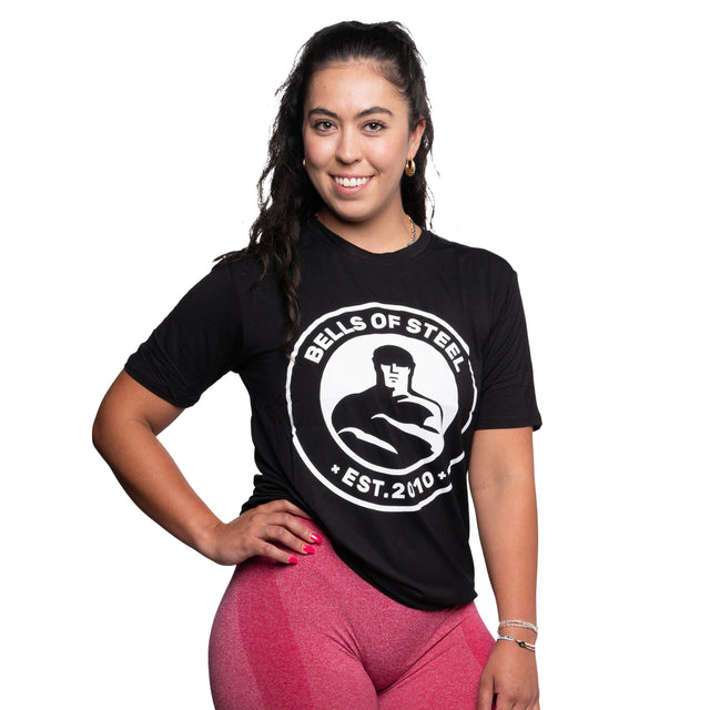 A person with long, dark hair is smiling at the camera while wearing a buttery soft bamboo workout shirt from Bells of Steel and pink leggings, standing against a white background. The shirt features the text "Bells of Steel EST. 2010.