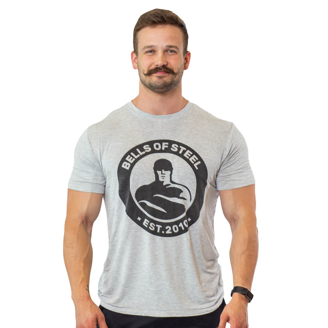 A man with a mustache is smiling while wearing a buttery soft Bamboo T-Shirt from Bells of Steel, adorned with the phrase "Bells of Steel Est. 2010" and a graphic of a muscular figure. He is standing against a plain white background.