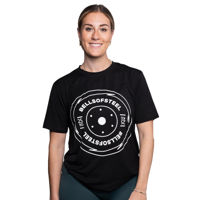 A person smiling and wearing a Bells of Steel Bamboo T-Shirt, crafted from a buttery soft bamboo blend, featuring a circular graphic with "BELLS OF STEEL" and "25 KG" in white text.