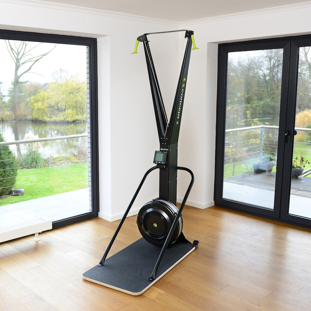 The Concept 2 SkiERG With PM5, created by Concept 2 for a comprehensive full-body workout, is placed in a bright room with large windows. Outside, the peaceful scene of trees, grass, and a pond enhances the atmosphere. Featuring black and green accents along with adjustable resistance on its compact digital screen, it serves as an excellent fitness companion.