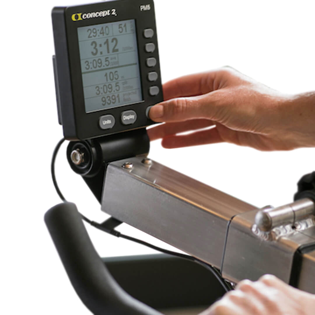 Close-up of a person's hand adjusting the PM5 Performance Monitor on a Concept 2 BikeERG. The screen displays workout stats such as time, distance, and pace. In the foreground, the handlebars complement this setup for an effective low-impact workout.