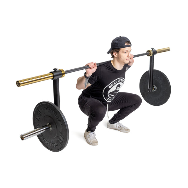 A person in a black t-shirt and cap performs a squat with the Camber Bar Attachment - Pair by Bells of Steel across their shoulders, relying on stabilizer muscles for balance. Large round weights are secured with barbell collars, while the plain white background keeps the focus on their form.