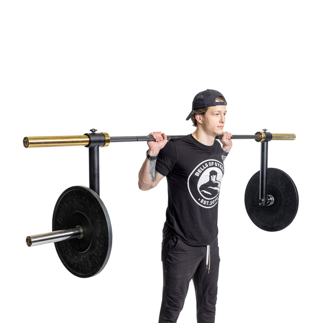 A person stands ready to lift a yoke barbell equipped with Bells of Steel's Camber Bar Attachments, which are holding the large weights in place. They are dressed in a black cap, T-shirt, and pants against a white background.