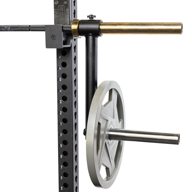 Close-up of a gym squat rack showcasing the Camber Bar Attachment - Pair by Bells of Steel, featuring a numbered vertical beam with weight plate storage. Two metal bars hold a gray weight plate with barbell collars and extend horizontally from the beam, providing stability and safety during exercises.