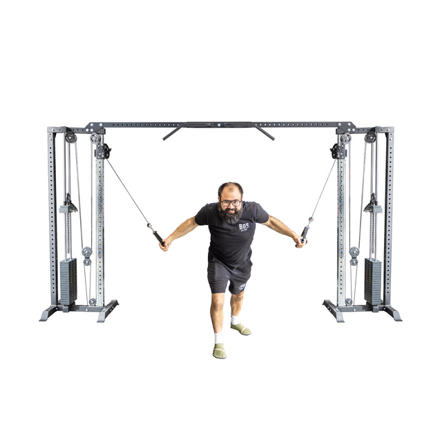 A man wearing glasses exercises with a Bells of Steel Cable Crossover Machine, pulling cables forward while lunging. He's in a black T-shirt, matching shorts, and gray socks against a white background.