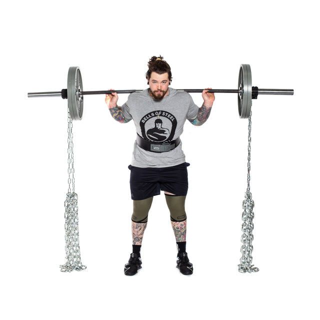 A tattooed individual holds a barbell across their shoulders, equipped with Bells of Steel Weightlifting Chains and weights for progressive resistance. They are dressed in a gray T-shirt, black shorts, and leggings, all set against a plain white background.