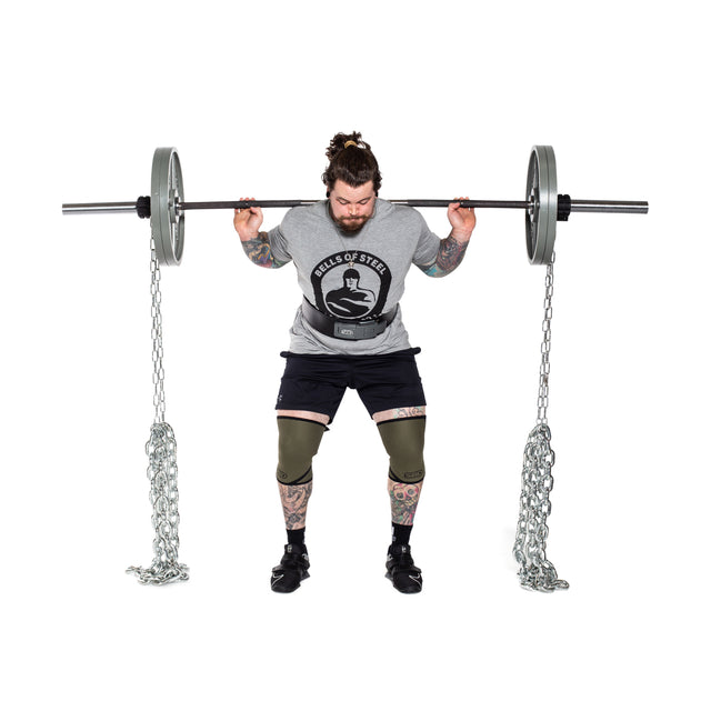 A tattooed individual with a bun stands in front of a white background, preparing to squat with a barbell enhanced by Bells of Steel Weightlifting Chains hanging from each end. Dressed in a gray shirt, black shorts, and knee sleeves, they focus on achieving progressive resistance.