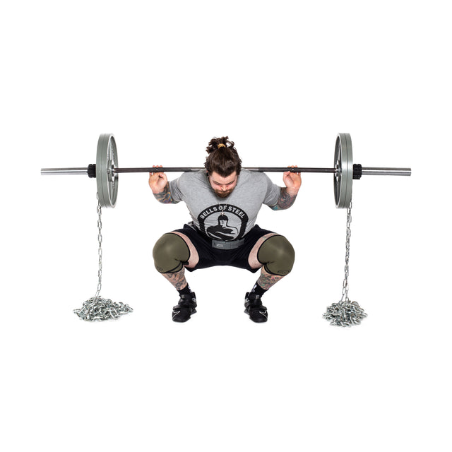 A muscular individual with tattoos incorporates Bells of Steel Weightlifting Chains over a barbell as they perform squats. Dressed in a gray shirt, black shorts, and knee sleeves, their hair is tied back in a bun while concentrating on maximizing muscular stimulation during the workout.