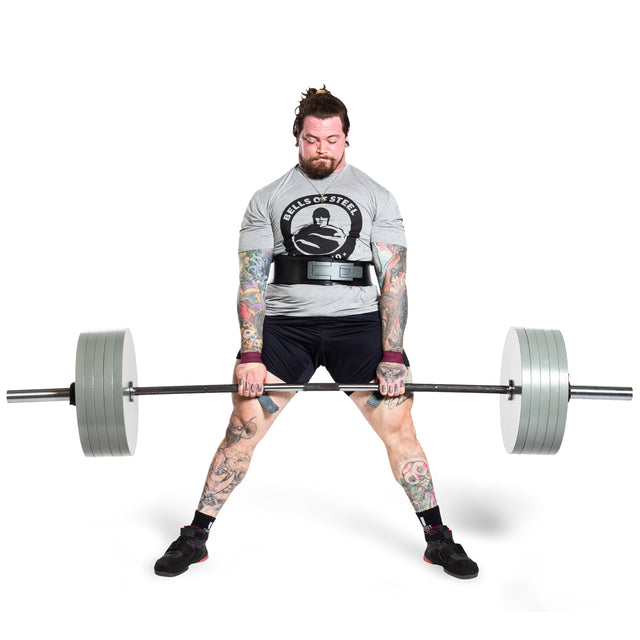 A tattooed individual is executing a deadlift using Bells of Steel Machined Iron Olympic Weight Plates on the barbell. Clad in a gray T-shirt, shorts, and a weightlifting belt, they concentrate intently on their lift.