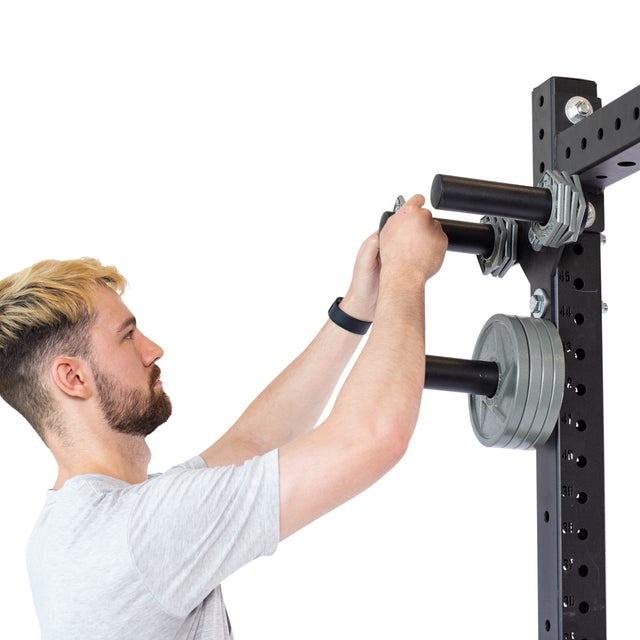 A short-haired person in a light gray T-shirt and black fitness tracker adjusts weights on a power rack. The Bells of Steel Change Plate Storage Pegs are seen with securely mounted plates, and the rack has multiple holes for adjustments.