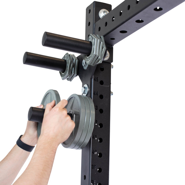 Two hands adjust weight plates on Bells of Steel’s Change Plate Storage Pegs, designed like a power rack. The black metal frame features horizontal pegs for securely holding weights, with gray plates standing out against the plain white background.